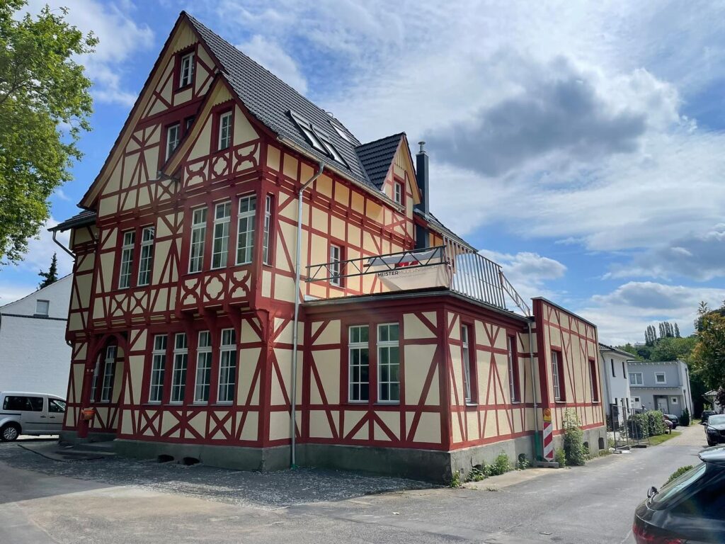 Ein traditionelles Fachwerkhaus mit roten Holzbalken und cremefarbenen Wänden steht in einer ruhigen Straße unter einem teilweise bewölkten Himmel. Mehrere Autos sind in der Nähe geparkt, und im Hintergrund sind kleine moderne Häuser zu sehen.