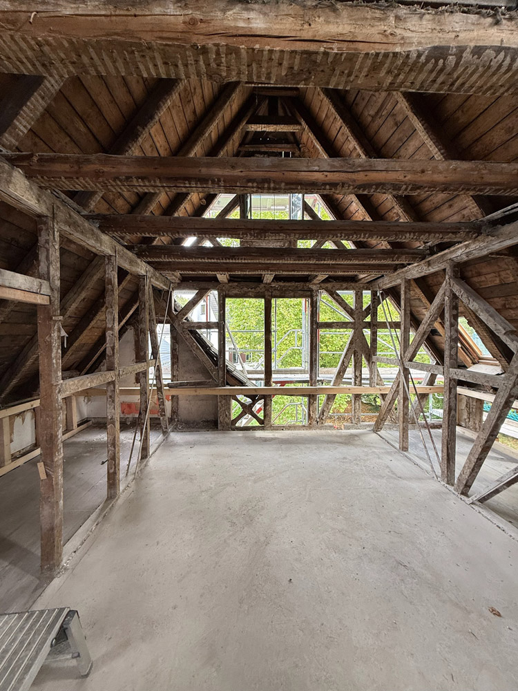 Ein Dachgeschoss mit freiliegenden Holzbalken, unfertigen Wänden und einem Betonboden. Große Fenster im hinteren Teil lassen natürliches Licht herein und geben den Blick auf das Grün draußen frei. Die Struktur scheint sich in der Renovierung oder im Bau zu befinden.