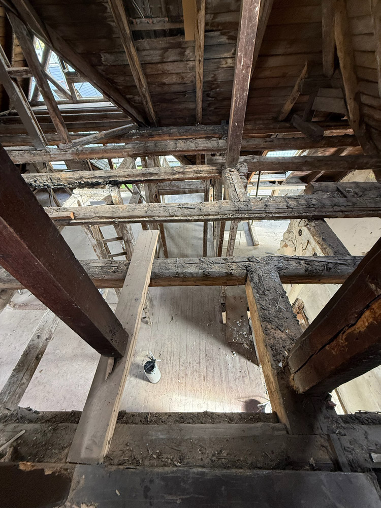 Blick von oben ins Innere eines Gebäudes, das gerade renoviert wird, mit freiliegenden Holzbalken, Dachsparren und einem teilweise offenen Fußboden mit Schutt darunter. Ein Eimer steht auf der unteren Etage.