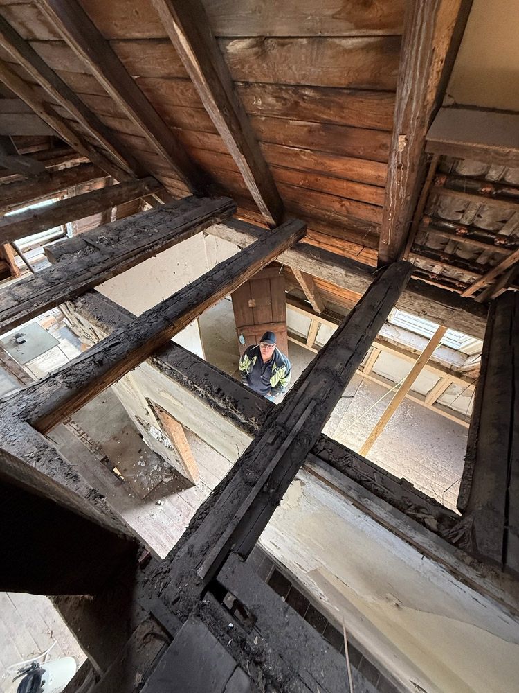 Eine Person, die einen Schutzhelm und eine Sicherheitsweste trägt, steht im unteren Stockwerk eines Gebäudes, das gerade renoviert wird, von oben gesehen durch freiliegende Holzbalken und eine unfertige Decke. Staub und Schutt sind sichtbar.