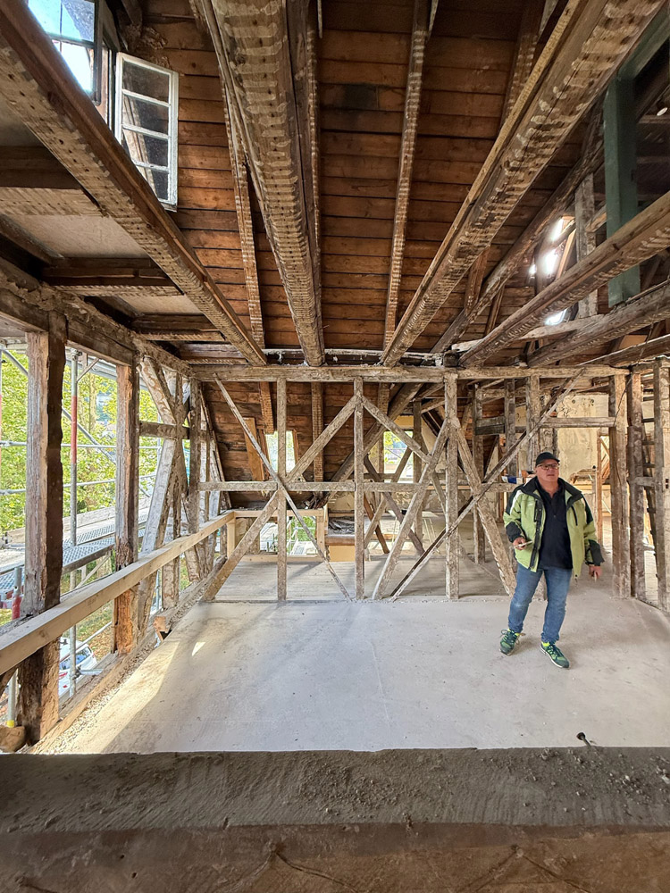 Eine Person in Jacke und Mütze steht in einem teilweise renovierten Gebäude mit freiliegenden Holzbalken, offenem Fachwerk und durch die Fenster einfallendem Sonnenlicht.