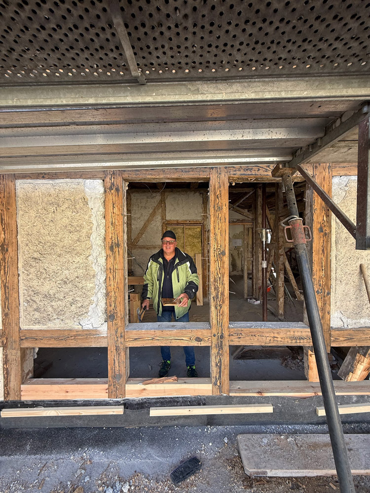 Eine Person in Arbeitskleidung und mit Mütze steht im Inneren eines teilweise renovierten Gebäudes, das von freiliegenden Holzbalken und grob verputzten Wänden eingerahmt wird. Baumaterialien und Werkzeuge sind überall auf der Baustelle zu sehen.