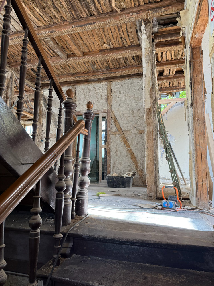 Ein altes, teilweise renoviertes Gebäudeinneres mit freiliegenden Holzbalken, einer abgenutzten Treppe mit Holzgeländer, Bauwerkzeugen und auf dem Boden verstreuten Trümmern. Sonnenlicht dringt durch Lücken in den Wänden und der Decke.
