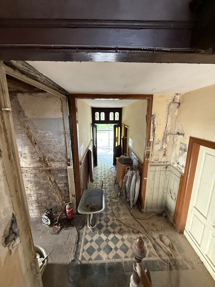 Ein Blick von einem Treppenhaus aus auf einen renovierten Flur mit freigelegtem Putz und Ziegeln, Bauwerkzeugen, einer Schubkarre, verhedderten Kabeln, einem Feuerlöscher und einer offenen Haustür, die Tageslicht hereinlässt.