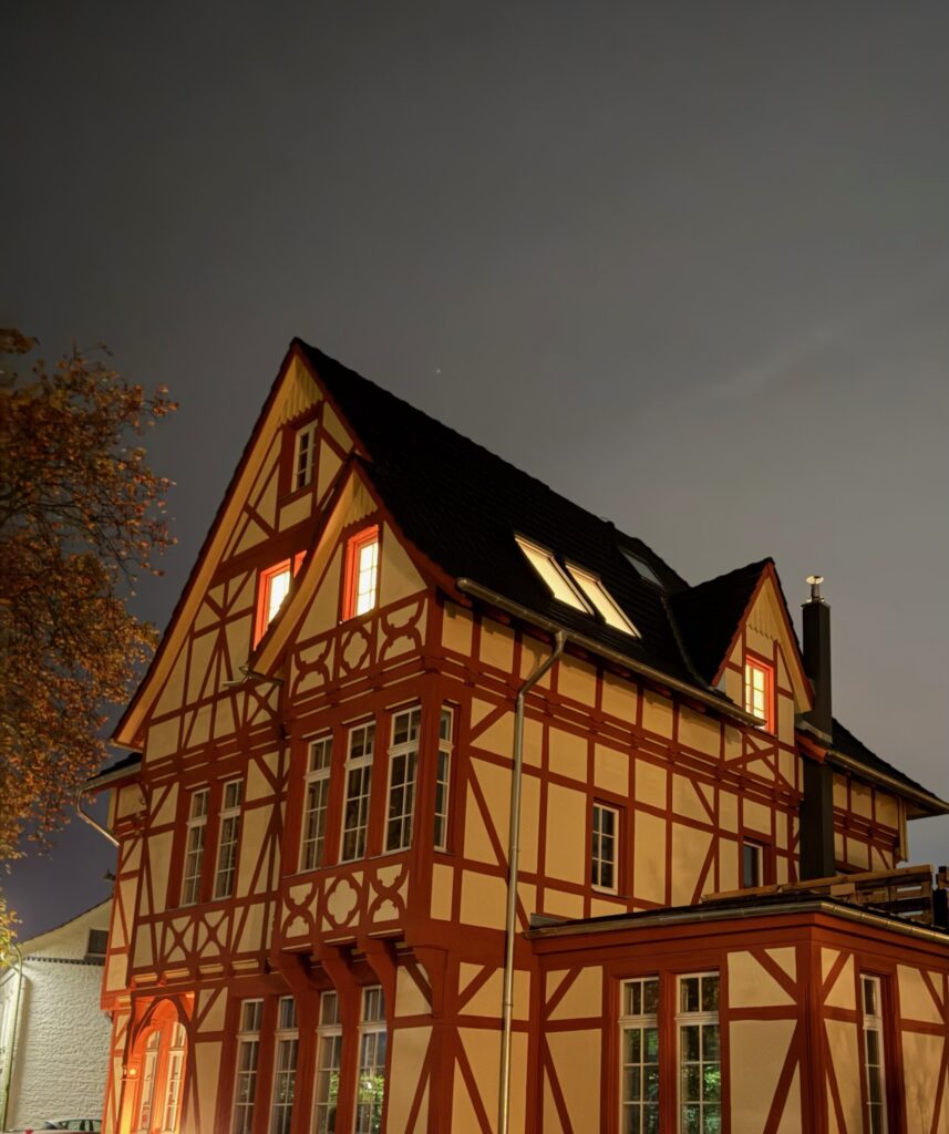 Ein traditionelles Fachwerkhaus mit roten und weißen Holzbalken ist nachts hell erleuchtet, vor einem wolkenverhangenen Himmel. Aus mehreren Fenstern leuchtet warmes Licht, und auf der linken Seite ist ein Baum zu sehen.