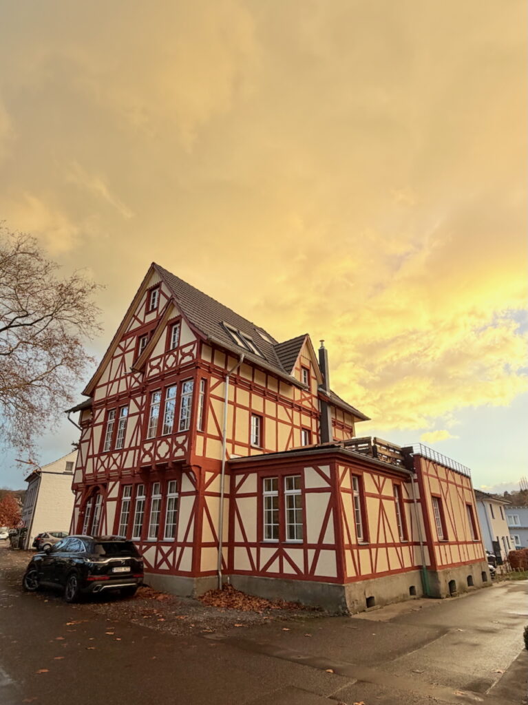 Ein traditionelles Fachwerkhaus mit roten Balken und cremefarbenen Wänden steht an einer Straßenecke unter einem dramatischen goldenen Himmel bei Sonnenuntergang, ein schwarzes Auto parkt in der Nähe und Herbstblätter liegen auf dem Boden.
