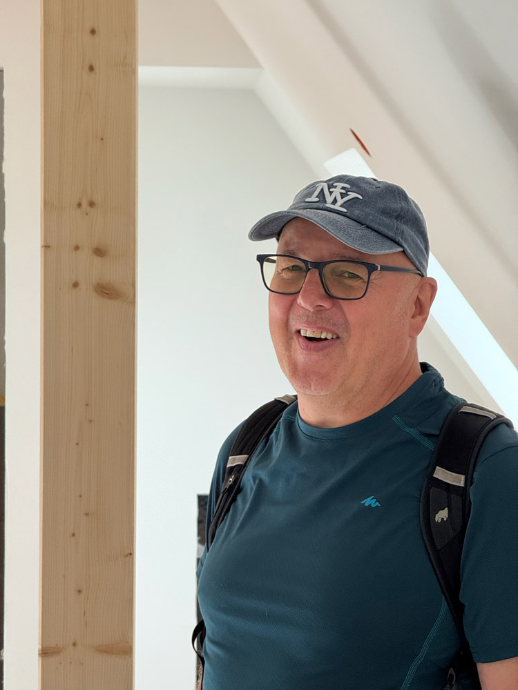 Ein lächelnder Mann, der eine Brille, eine blaue Baseballkappe und ein blaugrünes Hemd trägt und einen Rucksack mit sich führt, steht in einem Innenraum neben einem hellen Holzbalken und einer weißen Wand.