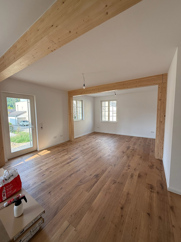Ein heller, leerer Raum mit großen Fenstern, hellen Holzböden, sichtbaren Holzbalken und weißen Wänden. Sonnenlicht strömt durch eine Glastür herein, und ein paar ausgepackte Gegenstände liegen in der Ecke auf dem Boden.
