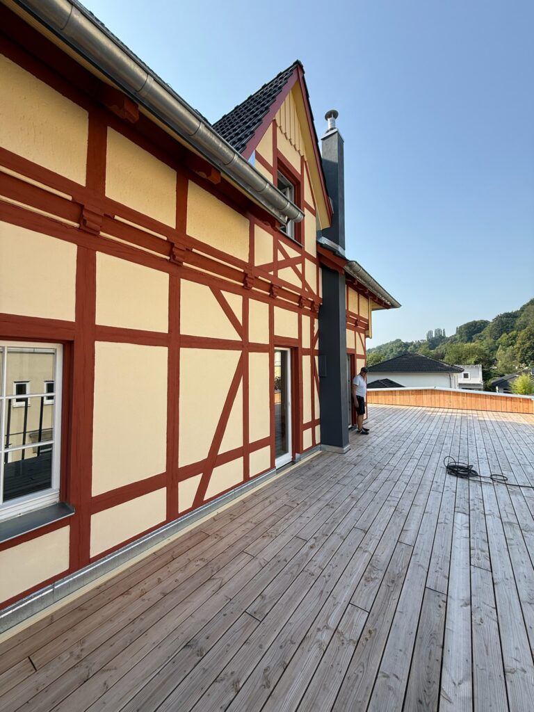 Eine breite Holzterrasse führt an einem traditionellen Fachwerkhaus mit roten und cremefarbenen Wänden entlang. Eine Person steht in der Nähe der Ecke, und im Hintergrund sind Bäume, Gebäude und ein klarer blauer Himmel zu sehen.