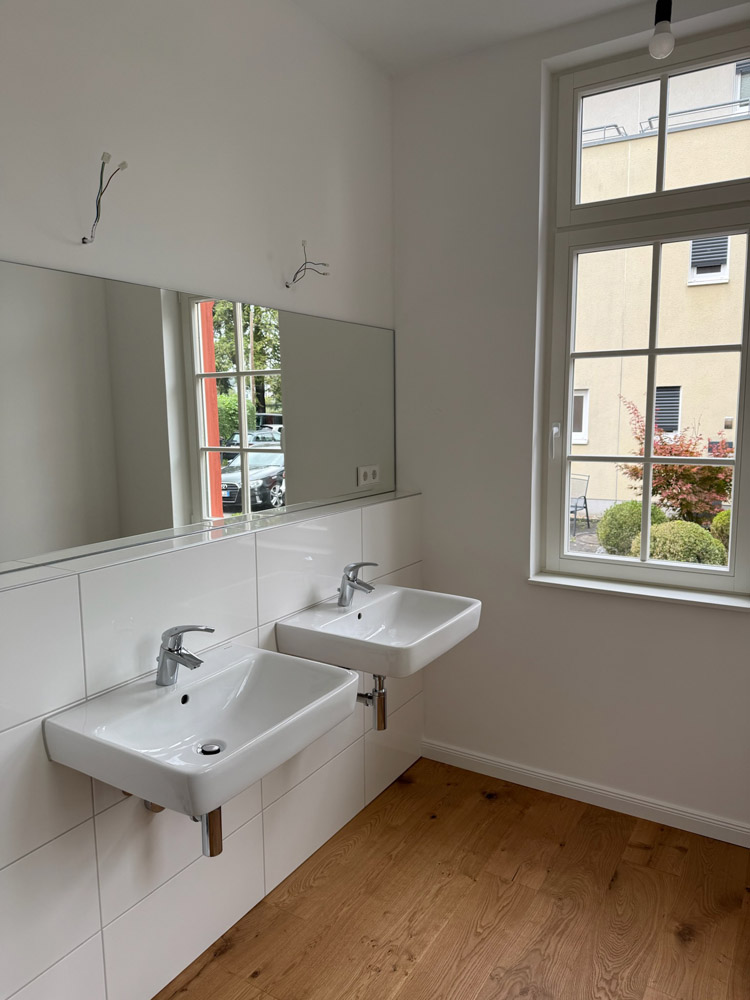 Zwei weiße Wandwaschbecken mit verchromten Armaturen sind unter einem großen Spiegel in einem hellen Badezimmer mit weißen Fliesenwänden, einem Holzboden und einem großen Fenster, das natürliches Licht hereinlässt, installiert.