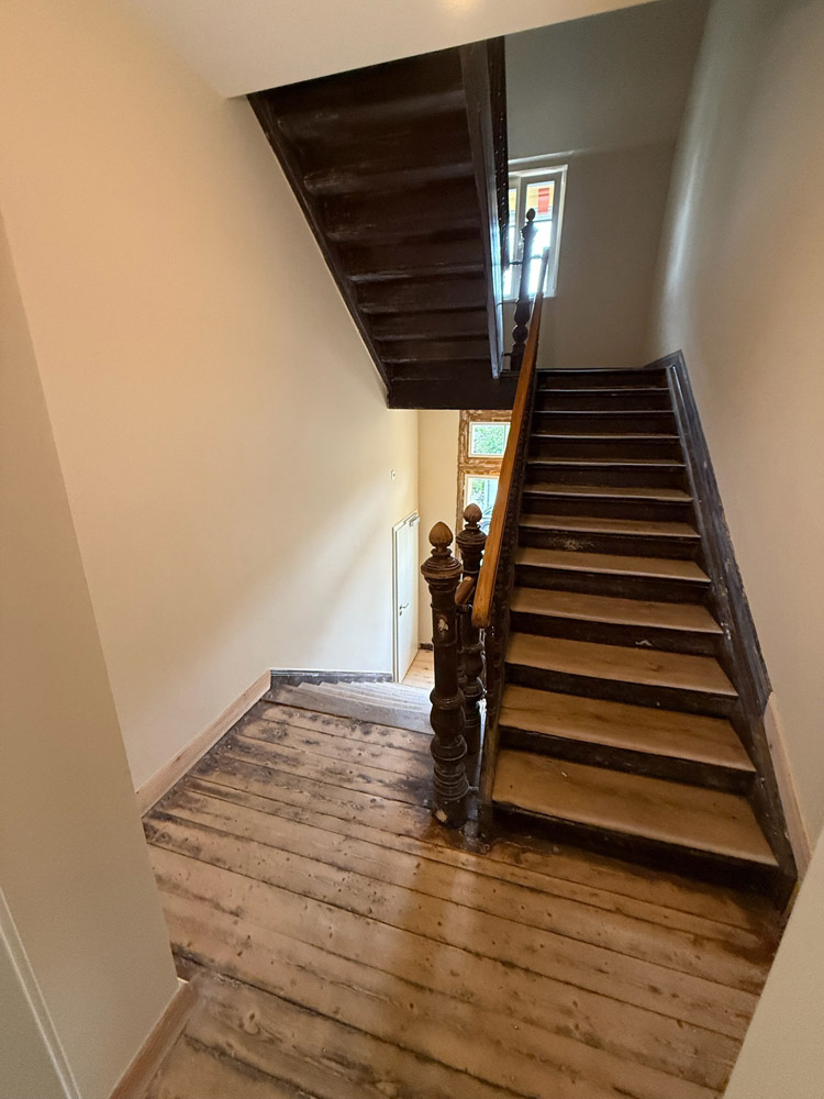 Eine Holztreppe mit dunklem Geländer biegt an einem Treppenabsatz ab und führt zu einem Fenster, das Tageslicht hereinlässt. Die Dielen und die Treppe wirken abgenutzt und unvollendet.