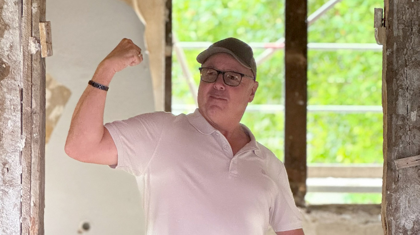 Ein Mann, der eine Brille, eine graue Mütze und ein hellrosa Polohemd trägt, beugt seinen rechten Arm und lächelt, während er in einem rustikalen, teilweise renovierten Raum steht, aus dem grünes Laub zu sehen ist.