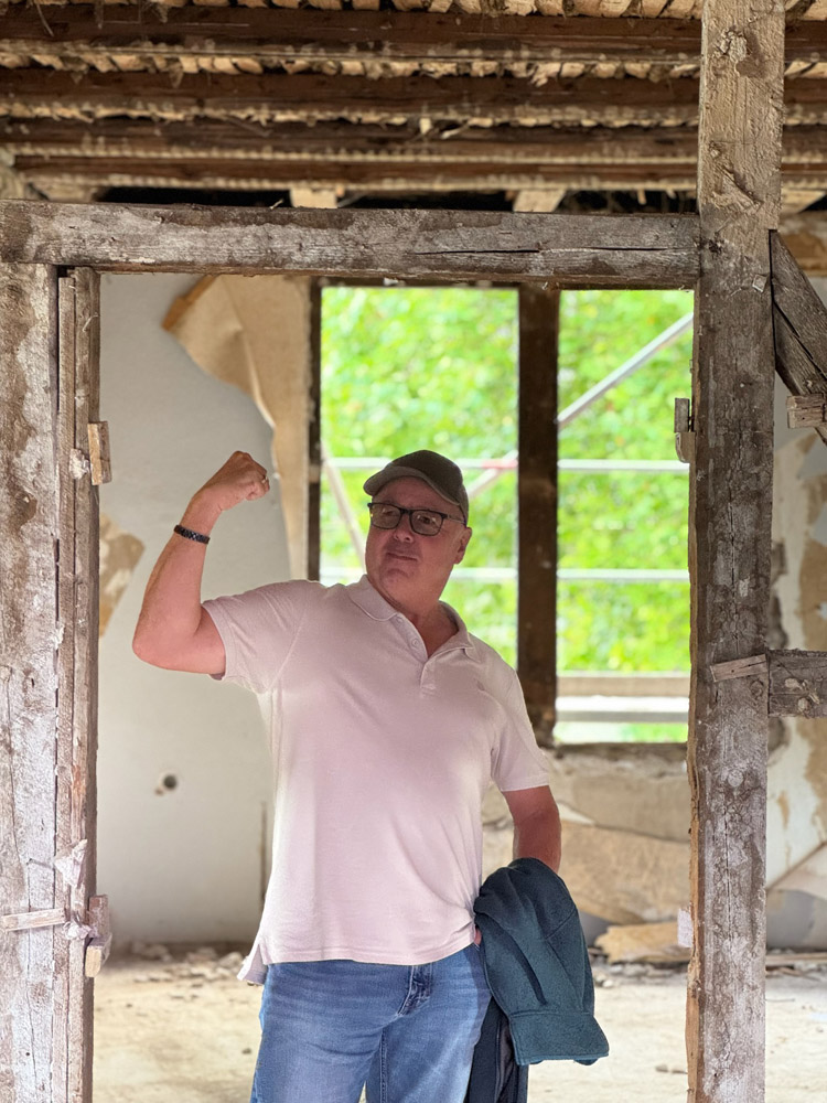 Ein Mann in einem weißen Polohemd und einer Mütze steht in einem abgenutzten, teilweise abgerissenen Gebäude, beugt seinen rechten Arm und hält eine Jacke in seiner linken Hand. Durch ein Fenster hinter ihm ist helles Grün zu sehen.