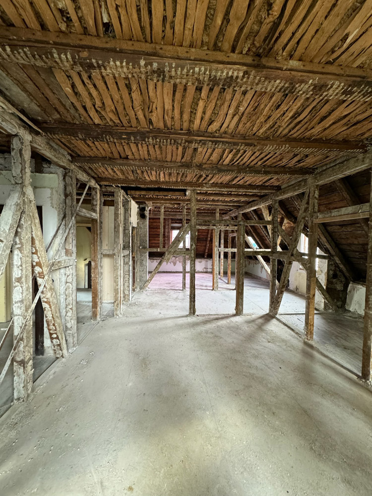 Ein leerer Dachboden mit freiliegenden Holzbalken und Latten an der Decke und den Wänden, unfertigen Fußböden und sichtbaren Stützen, die auf eine laufende Renovierung oder Konstruktion hindeuten.