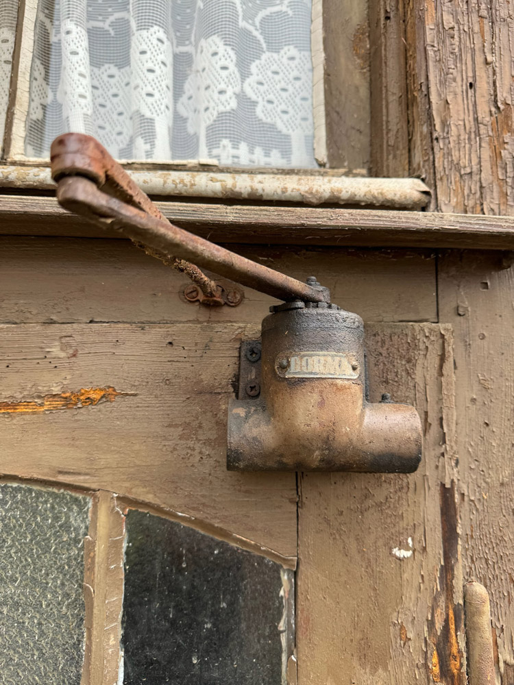Ein rostiger Metalltürschließer, der an einer alten Holztür mit abblätternder Farbe befestigt ist, neben einem Fenster mit Spitzenvorhängen und strukturierten Glasscheiben.