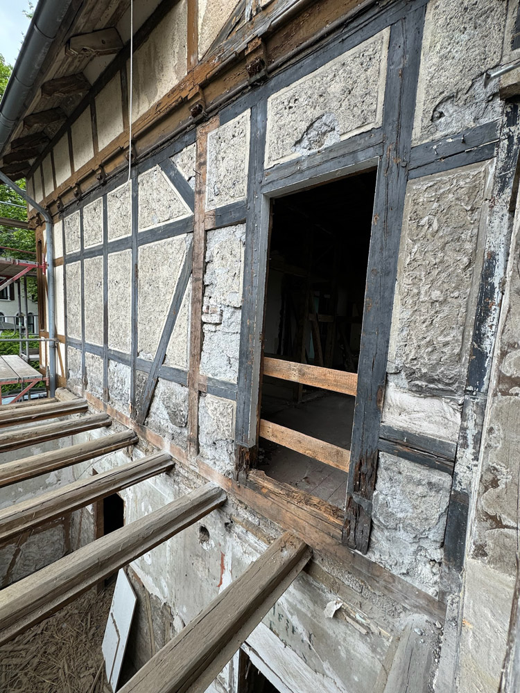 Ein teilweise restauriertes Fachwerkgebäude mit freiliegenden Holzbalken und fehlendem Fußboden, das eine offene Tür zeigt, die zu einem unvollendeten Innenraum führt. Außen sind Gerüste und Baumaterialien zu sehen.