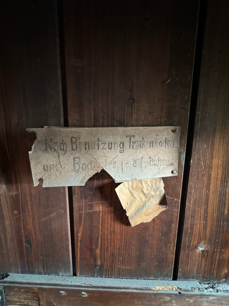 Ein verwittertes, zerrissenes Papierschild mit handgeschriebenem deutschen Text ist an eine dunkle Holzoberfläche gepinnt. Ein Teil des Schildes fehlt, wodurch ein Teil des Textes schwer zu lesen ist.