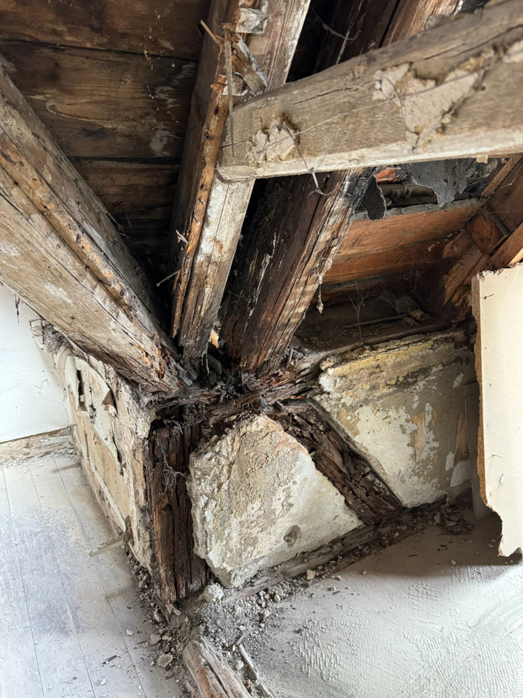 Freigelegte Holzbalken und Putz in einer Ecke einer maroden Wand und Decke, die erhebliche Wasserschäden, Fäulnis und Verfall aufweisen. Auf dem Boden sind Trümmer verstreut, die auf strukturelle Schäden und Vernachlässigung hinweisen.