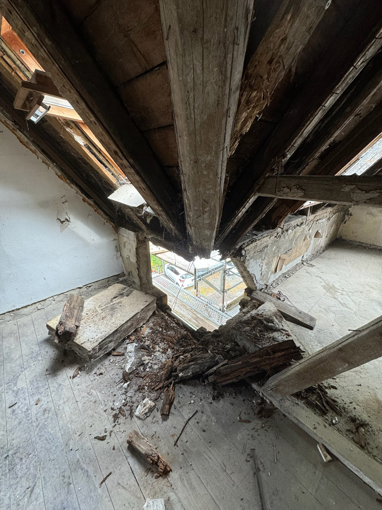 Blick aus dem Inneren eines alten Dachbodens mit einem großen, zerklüfteten Loch im Holzboden und im Dach, das Holzbalken und Trümmer freilegt und eine Straße mit geparkten Autos durch die Lücke zeigt.