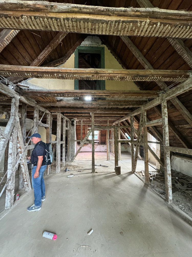 Ein Mann mit Baseballmütze und Rucksack steht in einem alten Dachgeschoss mit freiliegenden Holzbalken und einer schrägen Decke. Der Raum scheint unfertig zu sein, auf dem Boden liegen Schutt und Baumaterialien.