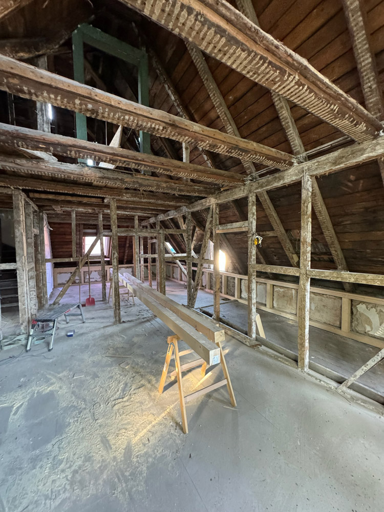 Ein renovierter Dachboden mit freiliegenden Holzbalken, entfernter Isolierung und Baumaterialien, darunter Sägeböcke und gestapeltes Holz, die auf dem staubigen Boden liegen. Durch kleine Fenster fällt natürliches Licht ein.