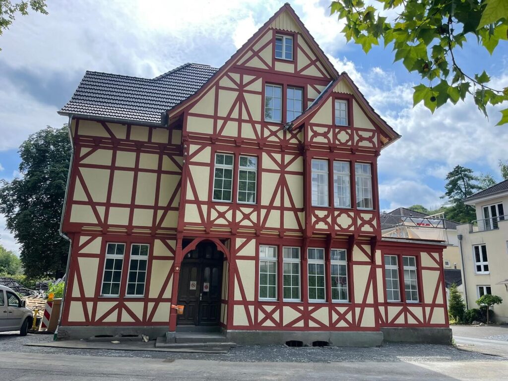 Ein großes, traditionelles Fachwerkhaus mit cremefarbenen Wänden und roten Holzbalken steht unter einem teilweise bewölkten Himmel. Das Gebäude hat mehrere Fenster und ein dreieckiges Dach. In der Nähe sind einige Bäume und ein weiteres Gebäude zu sehen.