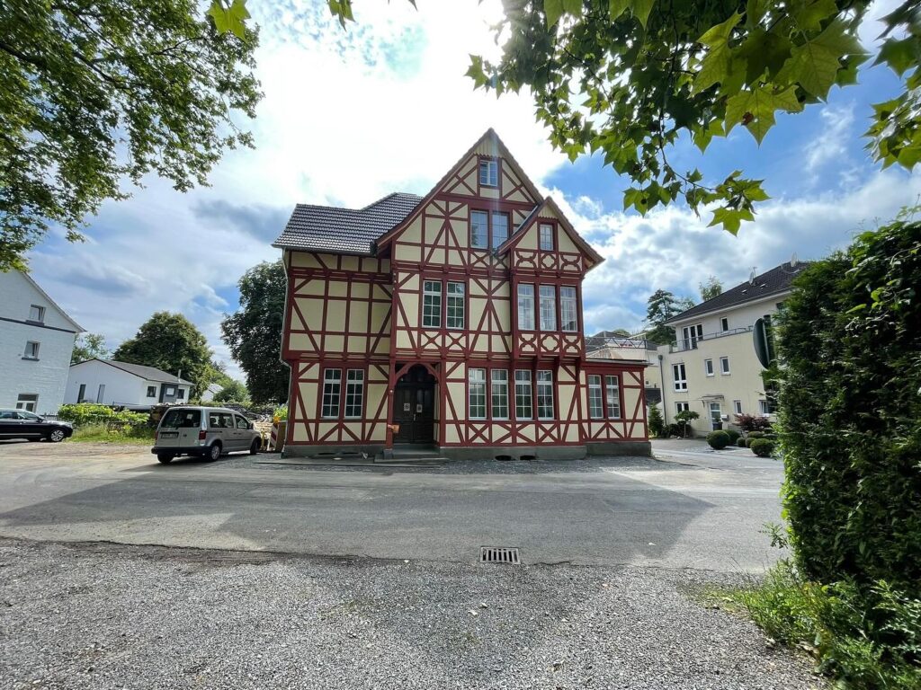 Ein traditionelles Fachwerkhaus mit roten Balken und cremefarbenen Wänden steht auf einem gepflasterten Platz, umgeben von Bäumen, Autos und Nachbargebäuden, über denen ein teilweise bewölkter Himmel hängt.